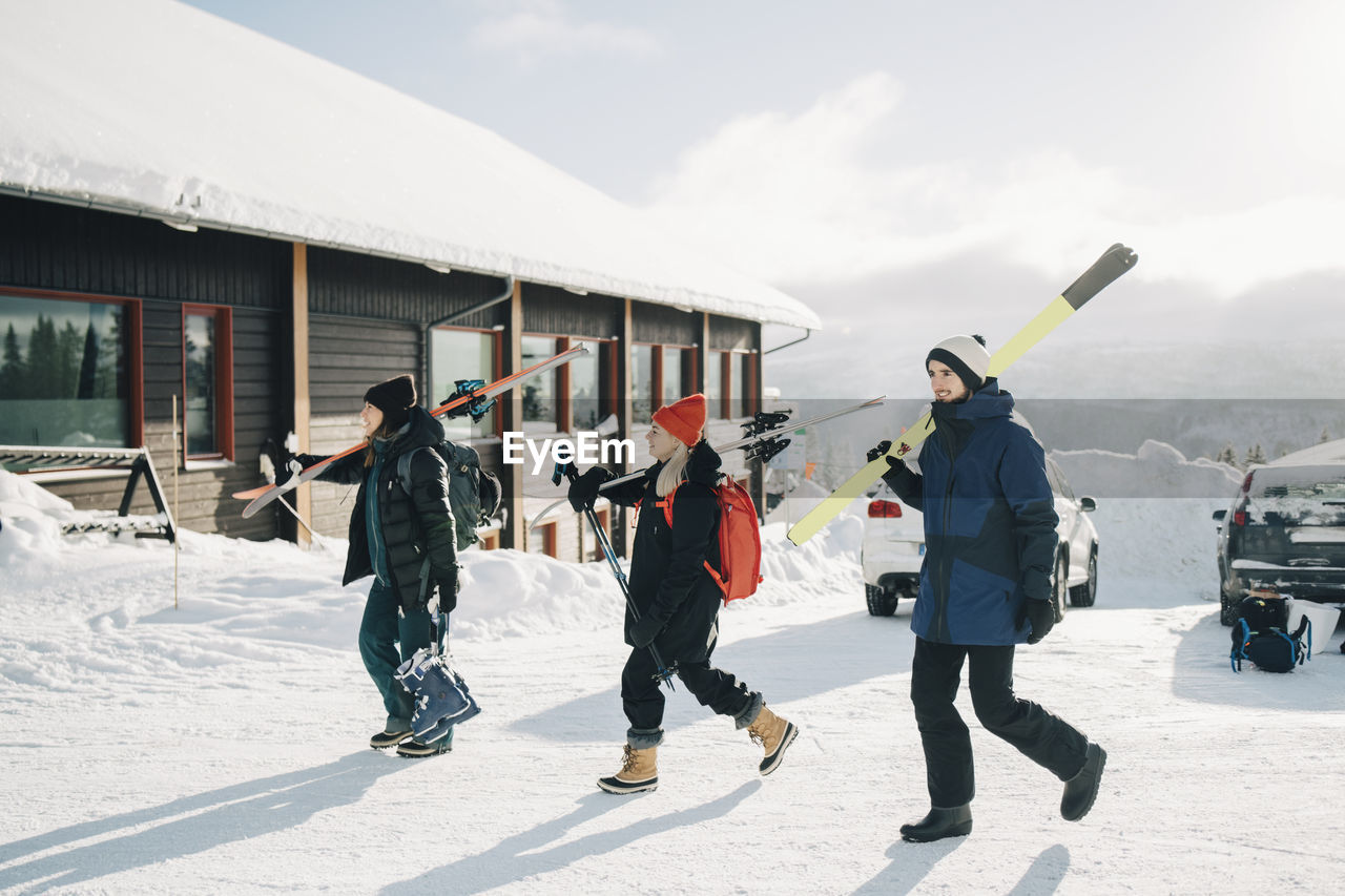 Full length of friends walking with skis at tourist resort during sunny day