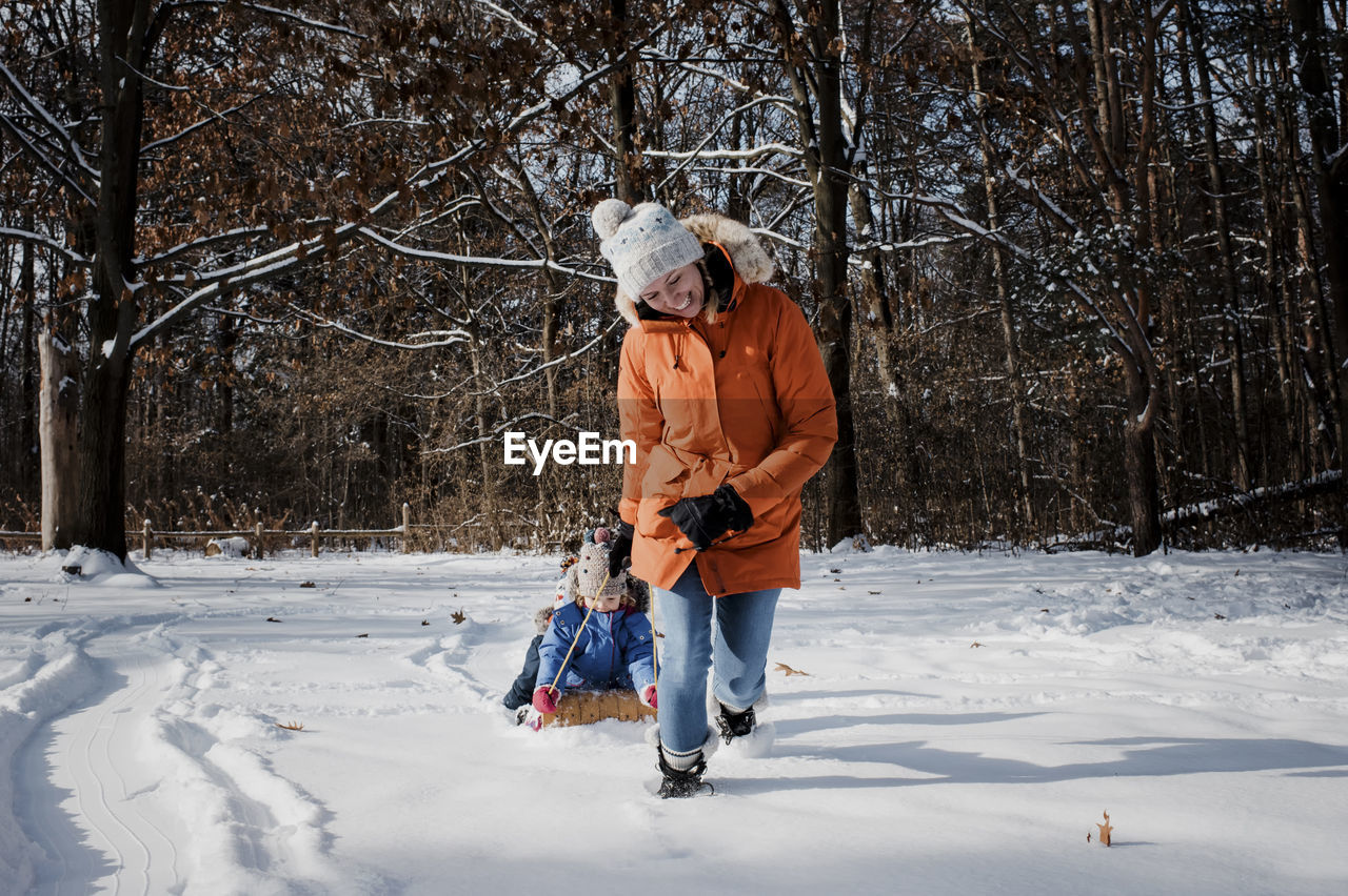 Happy mother pulling children sitting on sled in forest during winter