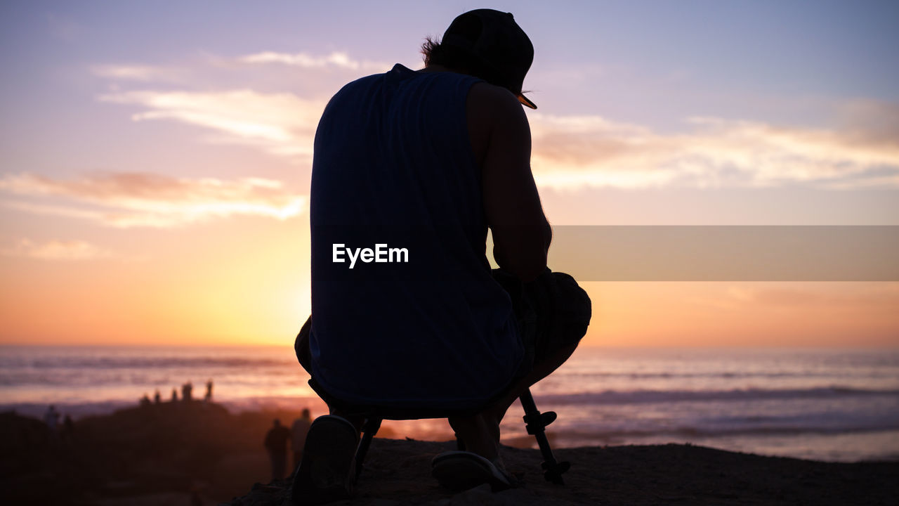 Silhouette photographer crouching with tripod at beach during sunset