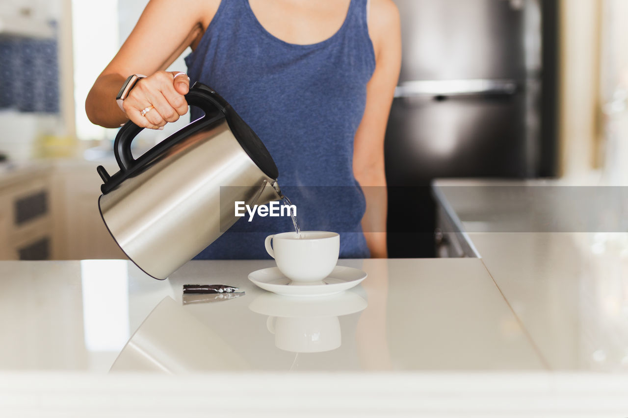 Woman pouring hot water from kettle in cup indoors.
