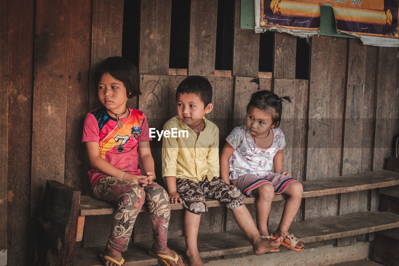 PORTRAIT OF SIBLINGS SITTING ON WOOD OUTDOORS