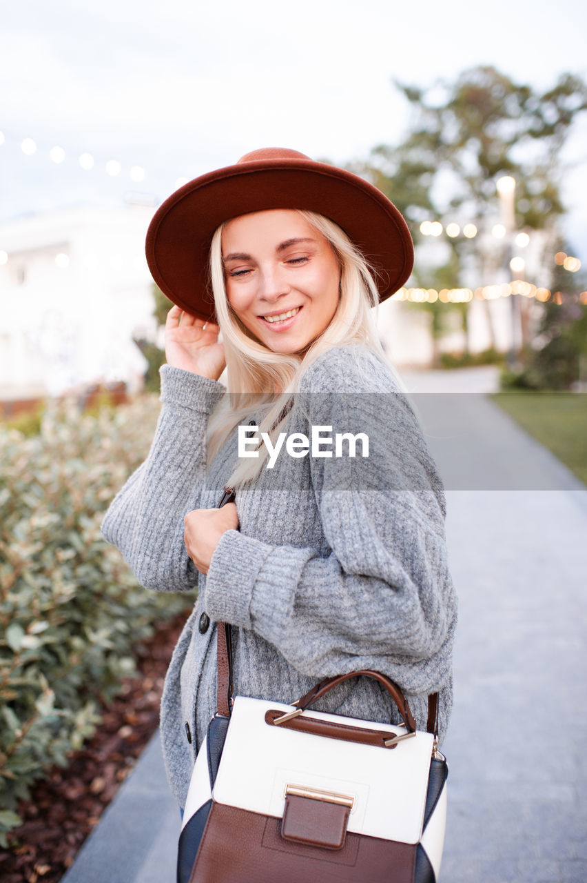 Laughing blonde cute young girl 20-24 year old wear hat, knitted wool sweater and holding trendy bag