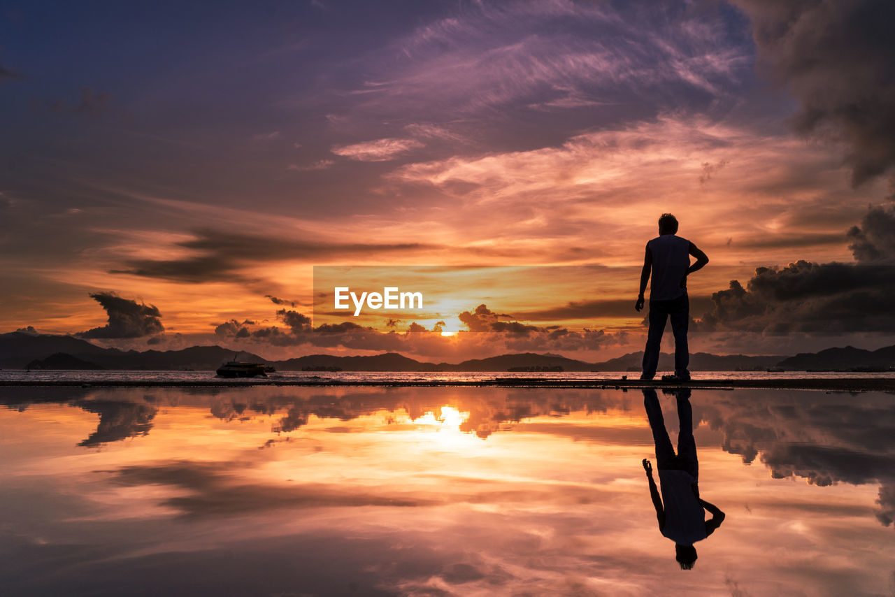 Rear view of man standing on lakeshore against sky during sunset