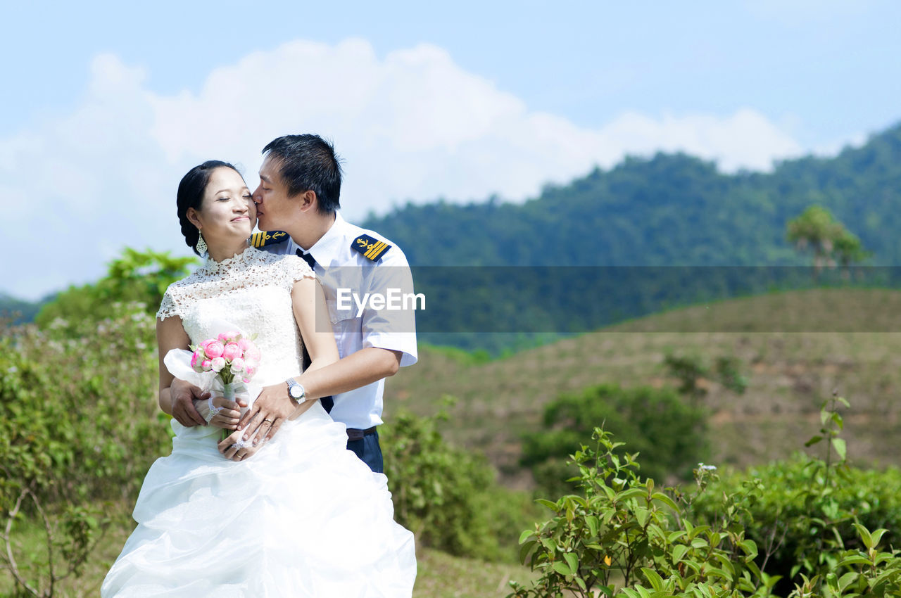 Happy bridegroom kissing bride on cheek against mountains