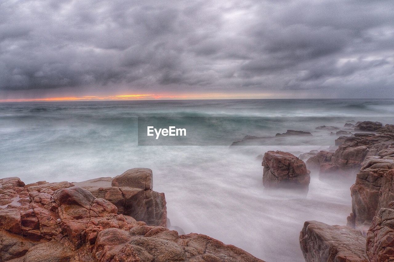 Rocks at sea shore against cloudy sky during sunset