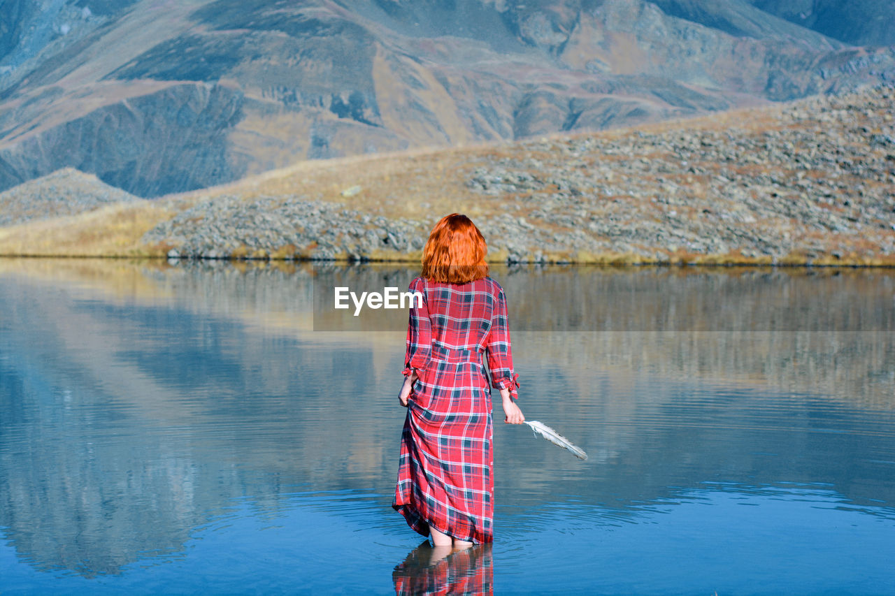 Rear view of woman holding feather while standing in lake by mountains
