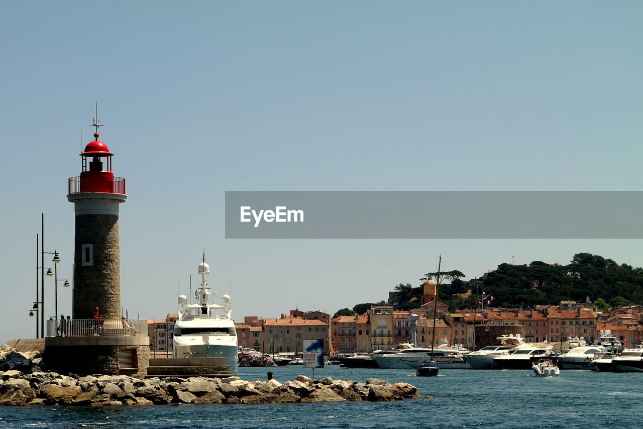 Lighthouse and buildings by boats moored at river against clear sky