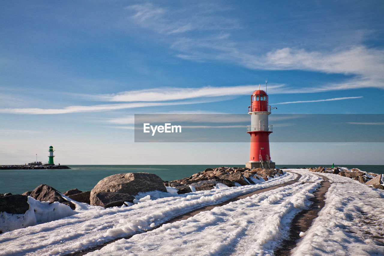 Snow covered pier towards lighthouse at sea against blue sky