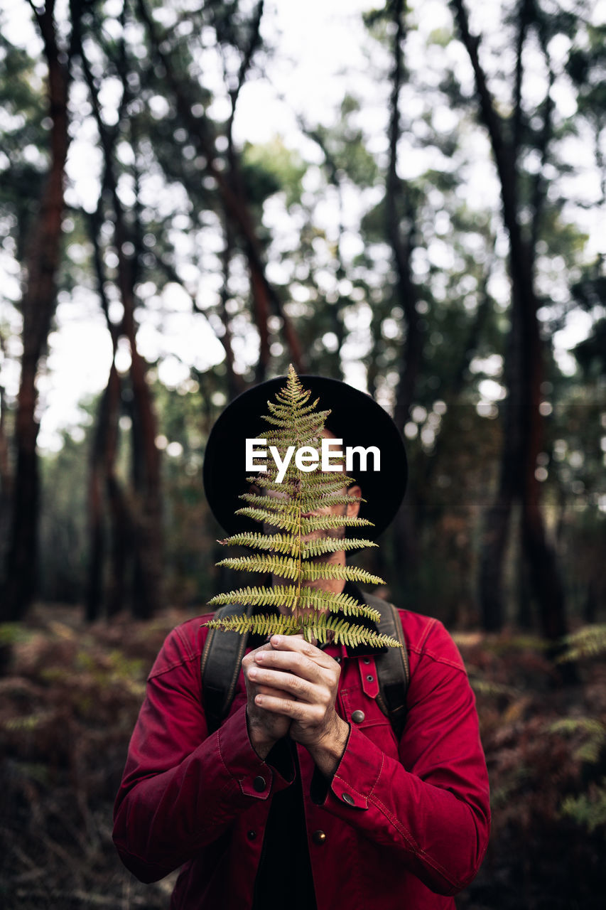 Adult male traveler in hat covering face with fern leaf standing against blurred trees of woods