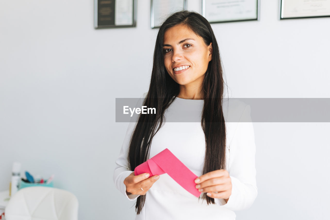 Young beautiful brunette woman with long hair cosmetologist holding tape ribbon in hands in office