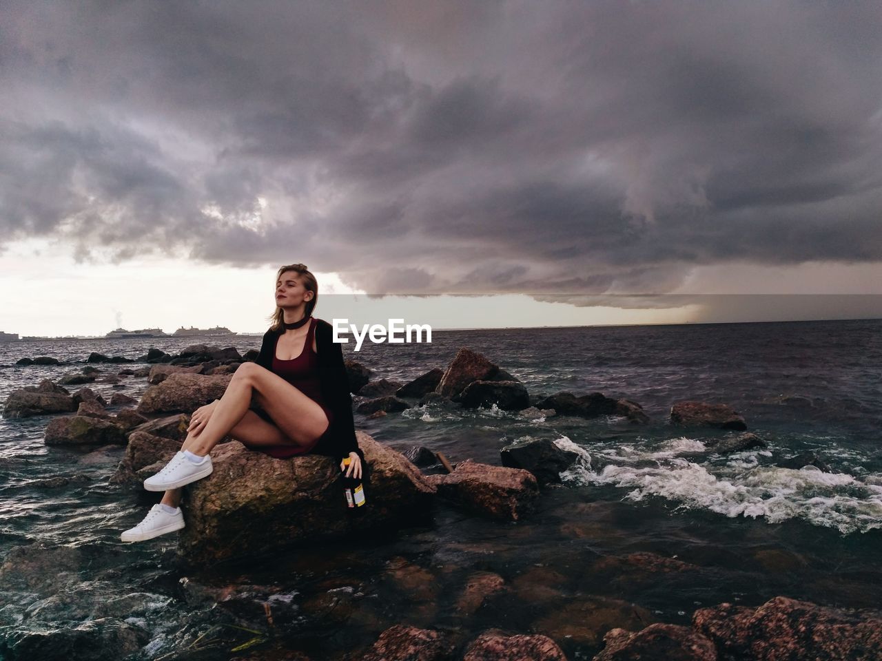 Beautiful fashion model sitting with liquor bottle on rock in sea against cloudy sky