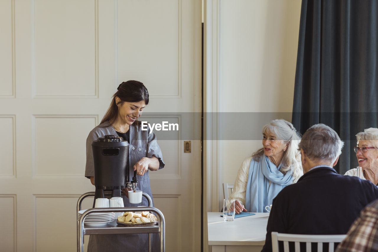 Smiling female healthcare worker serving coffee to senior women and man at retirement home