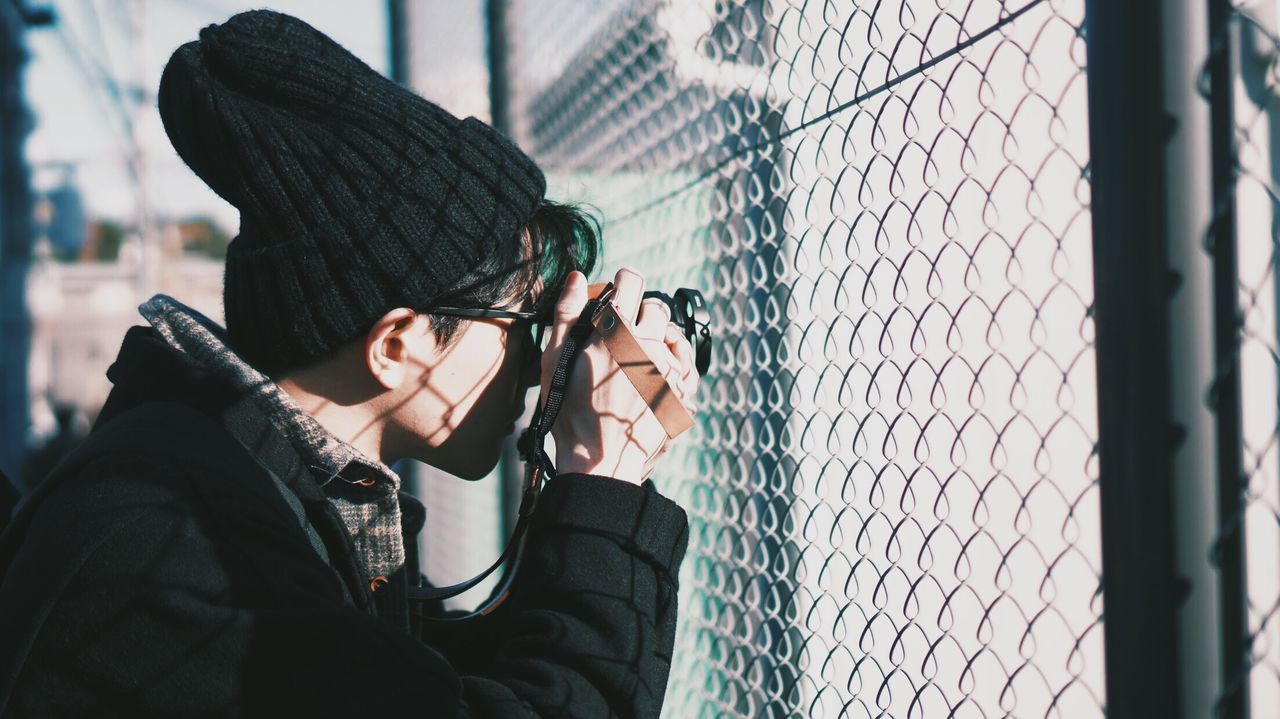 Side view of man photographing with camera by chainlink fence