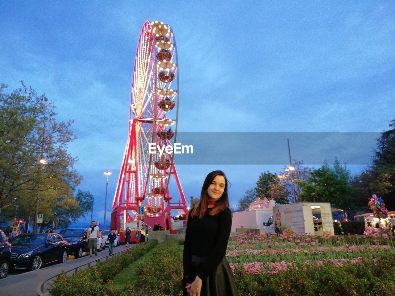 FULL LENGTH PORTRAIT OF SMILING YOUNG WOMAN AGAINST ILLUMINATED FERRIS WHEEL