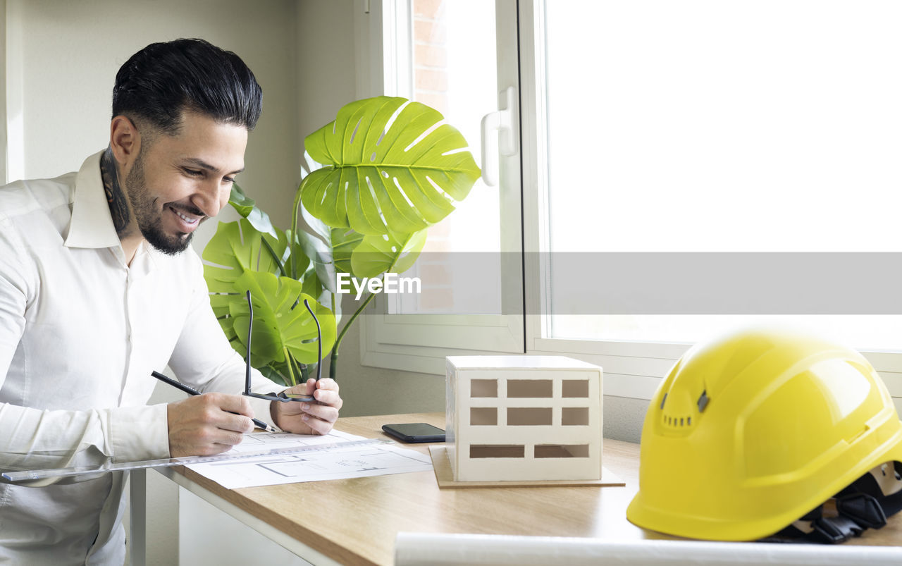 Smiling design professional holding eyeglasses while drawing blueprint at desk in office