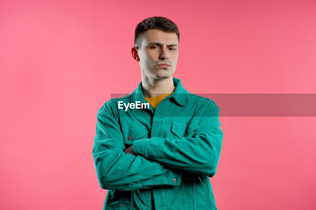 one person, studio shot, colored background, portrait, adult, young adult, waist up, indoors, looking at camera, standing, sleeve, outerwear, serious, front view, men, pink background, clothing, brown hair, copy space, casual clothing, photo shoot, contemplation, arms crossed, pink, hood, person, emotion, individuality, looking