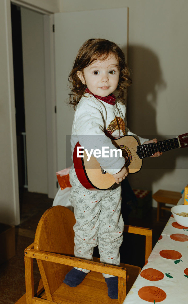 A little girl plays the guitar while standing on a chair at the table.