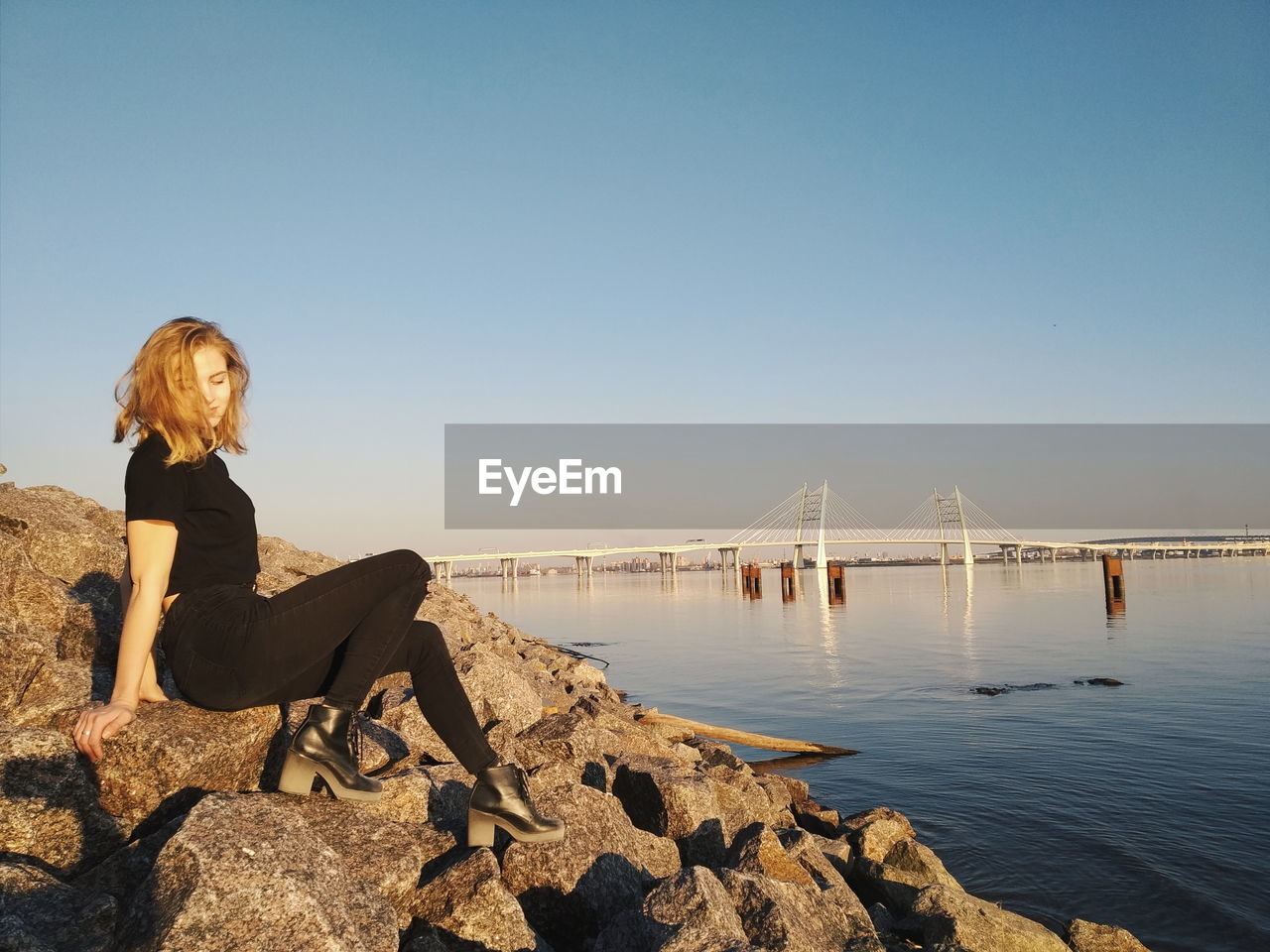 Full length of woman sitting on rocks by sea against blue sky