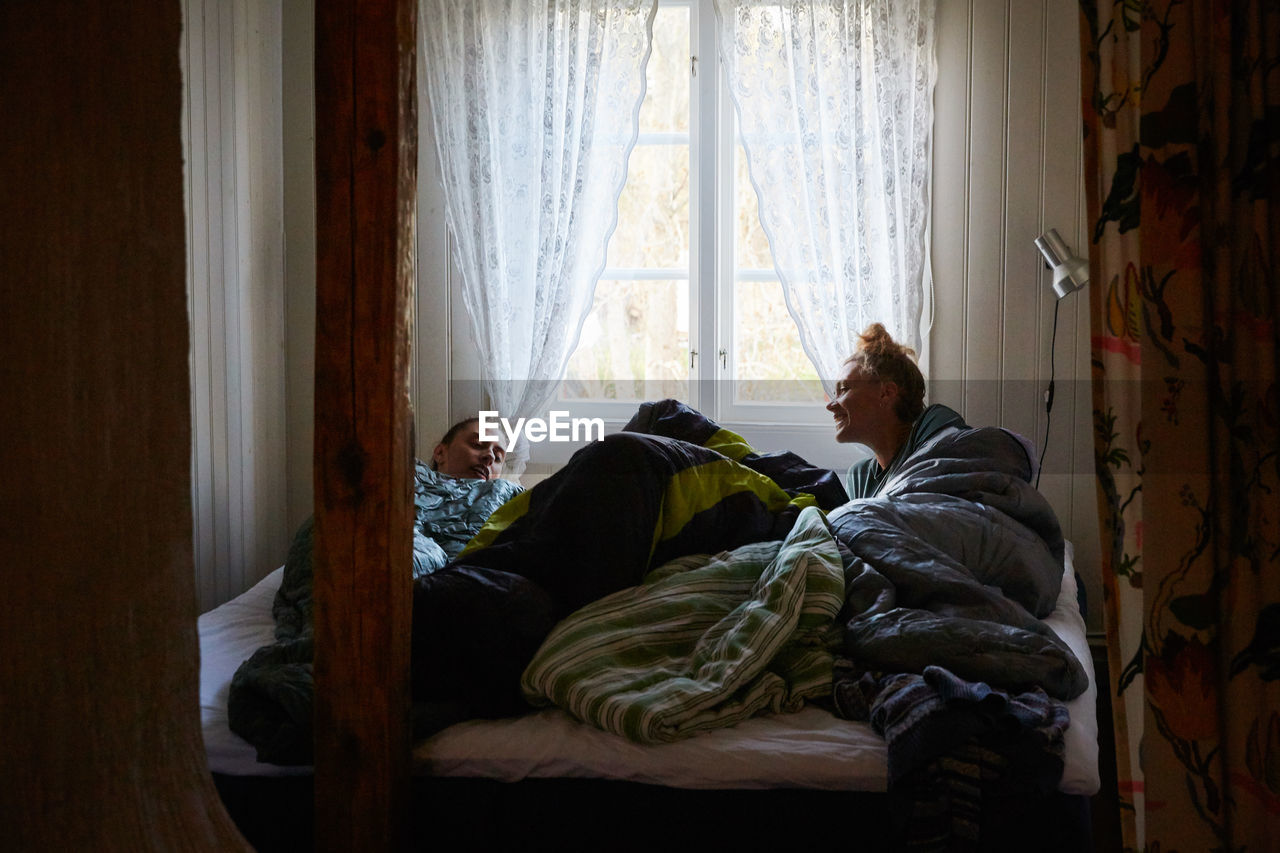 Male and female friends talking while relaxing on bed against window at home