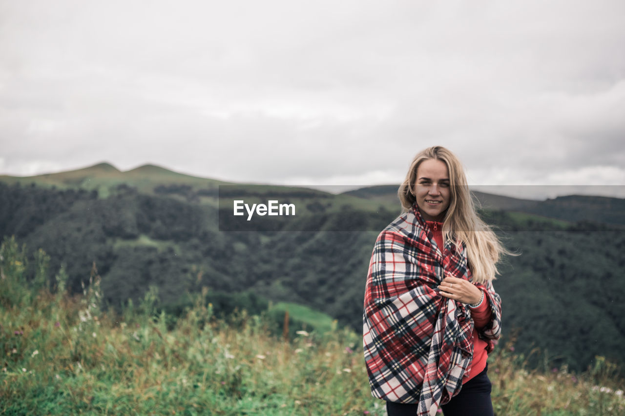 Portrait of smiling woman with blanket standing on mountain against sky