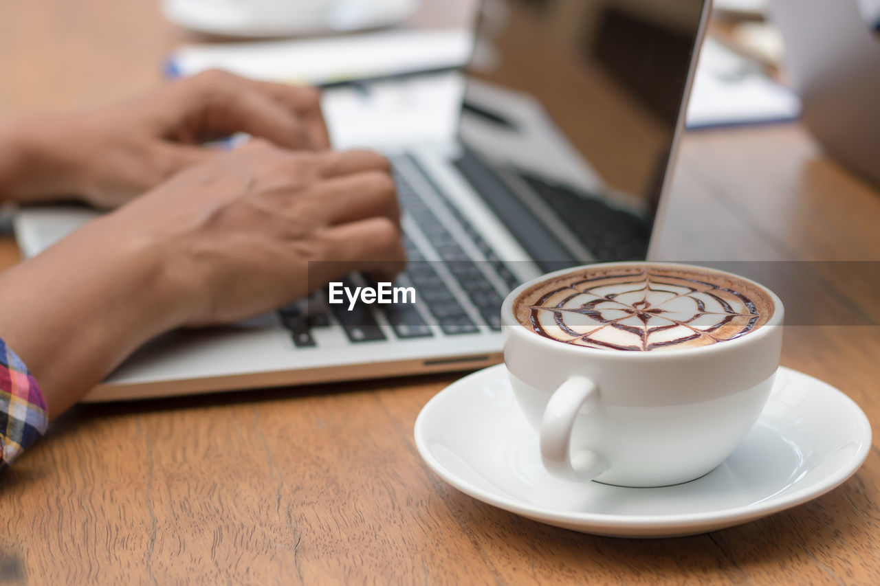 Cropped hands using laptop by coffee cup at table