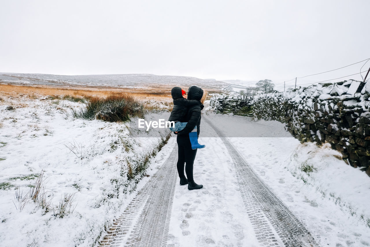 Full length of mother piggybacking son while standing on snow covered road during winter