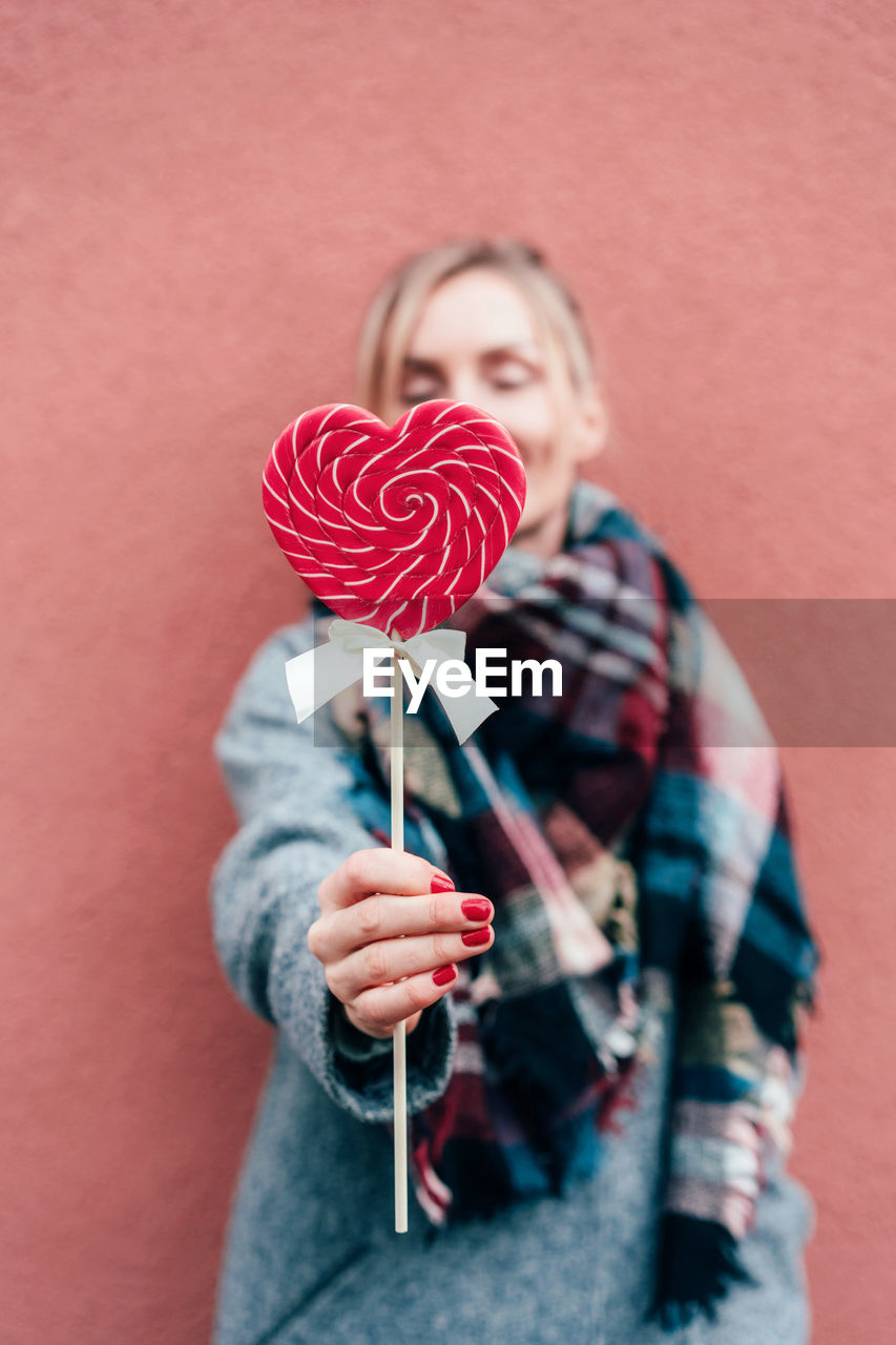 Woman holding heart shaped lollipop against wall