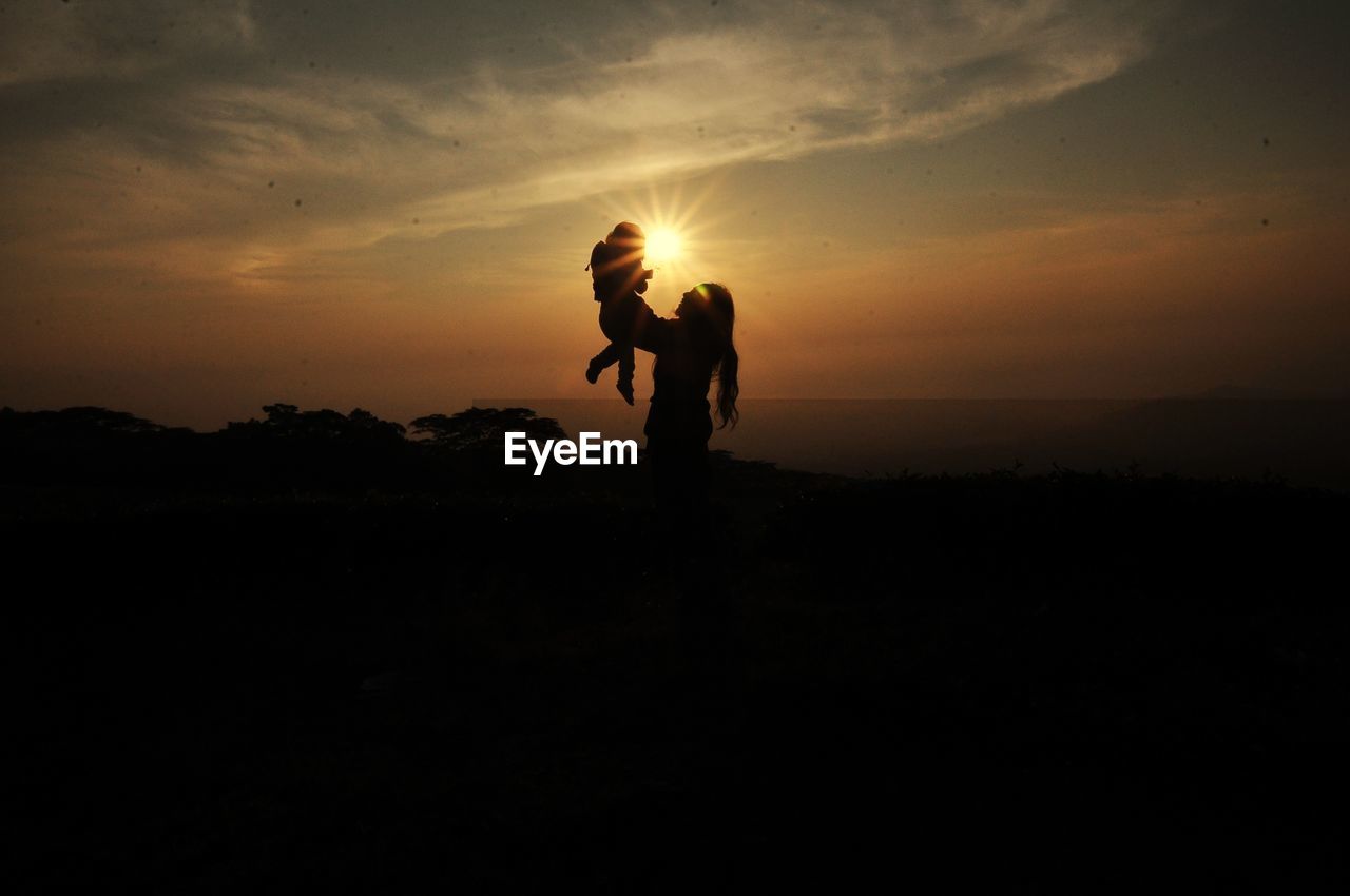 Silhouette woman picking up daughter against sky during sunset