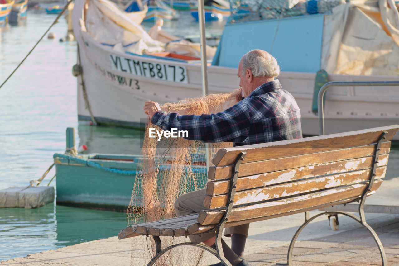 MAN SITTING ON BENCH BY BOAT