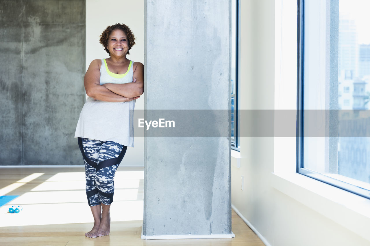 Happy woman with arms crossed looking away while leaning on architectural column in yoga studio