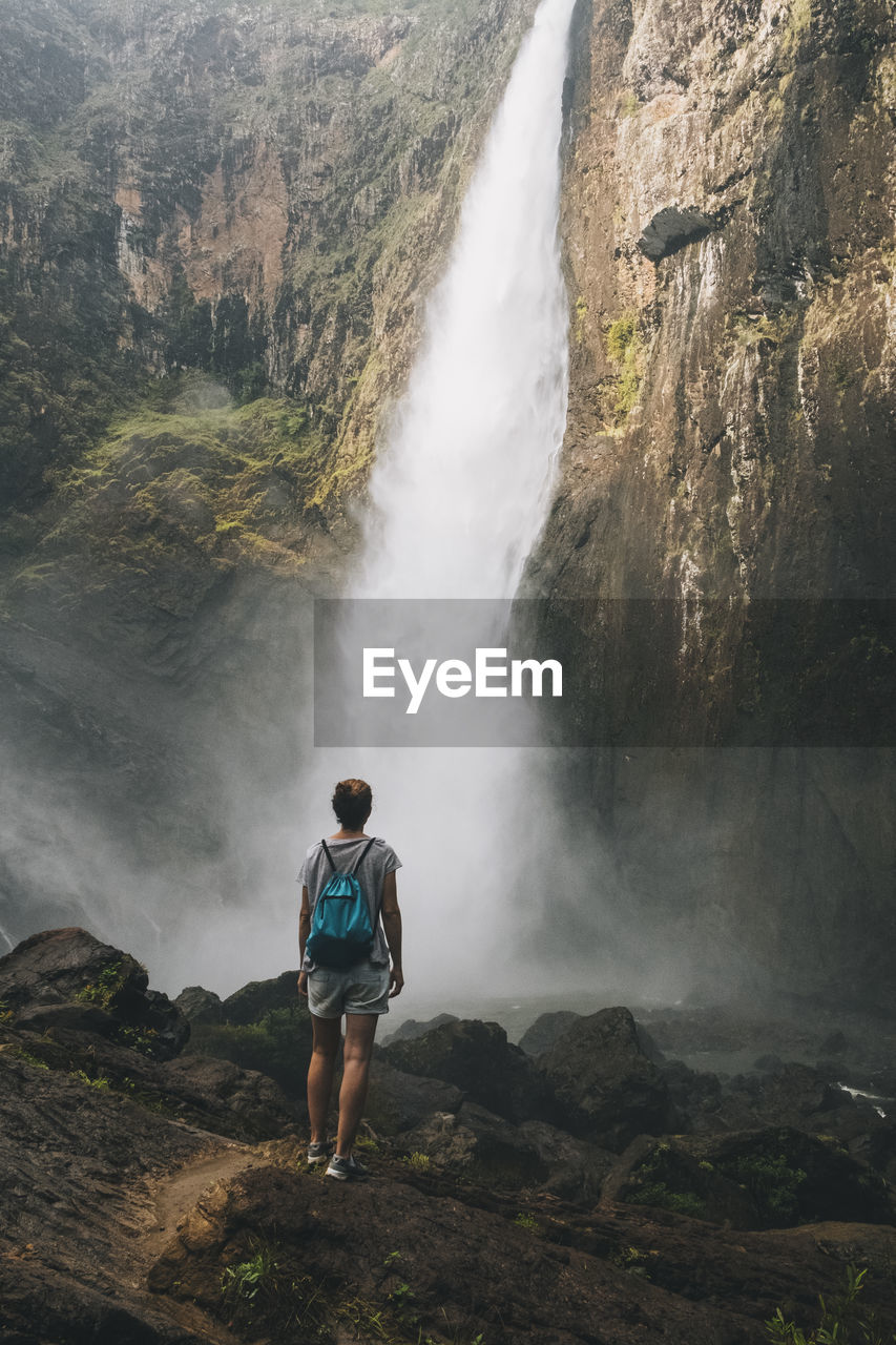 Rearview of a young woman looking at the waterfall while standing in a forest, queensland, australia.