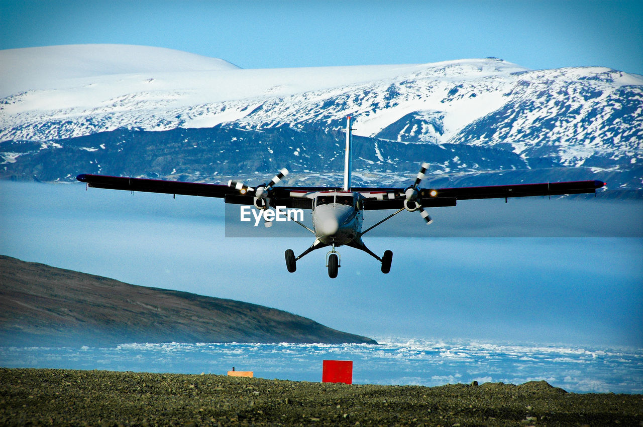 Biplane flying over lake against snowcapped mountains during sunny day