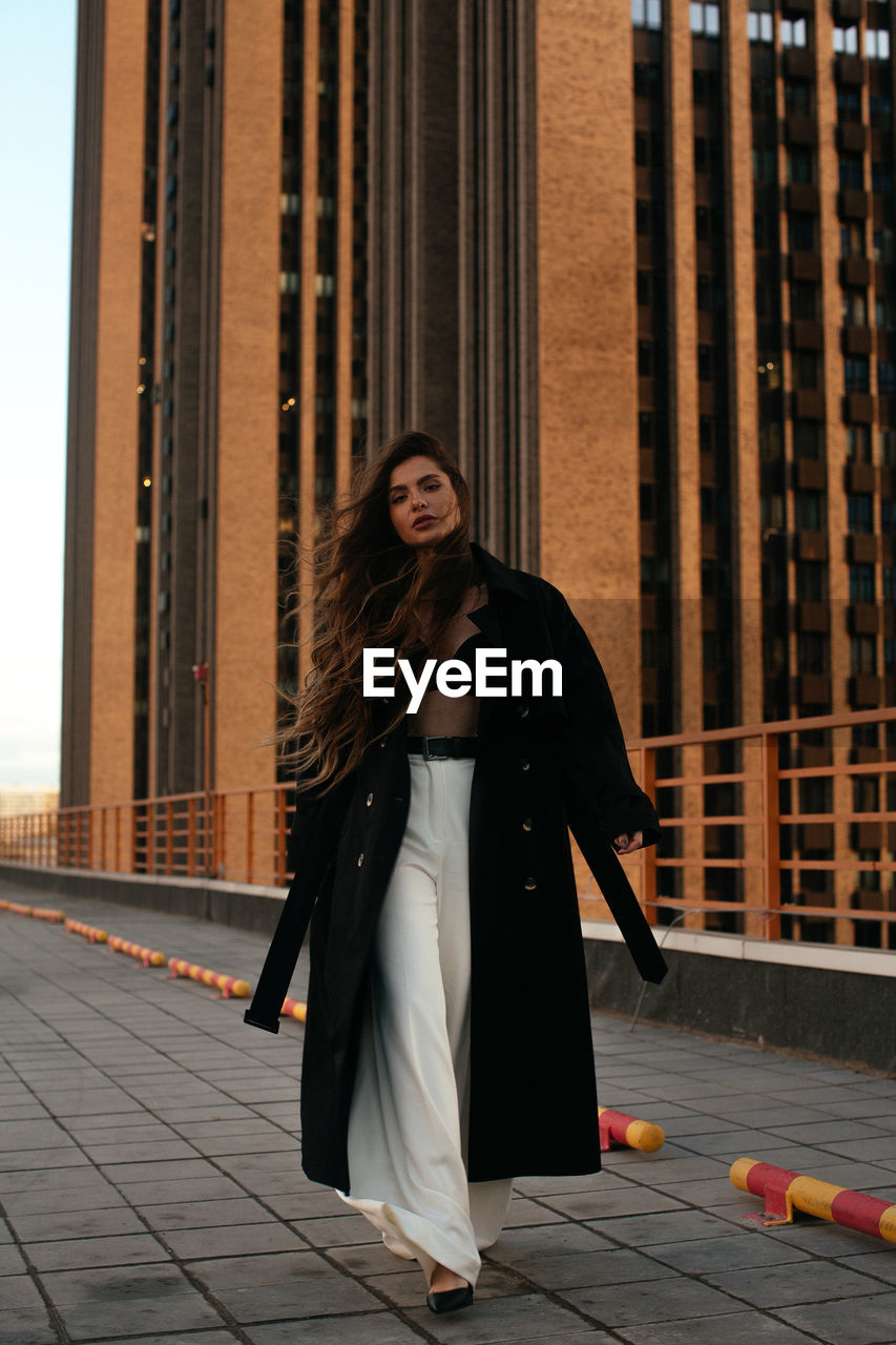 FULL LENGTH PORTRAIT OF WOMAN STANDING AGAINST CITY IN BACKGROUND