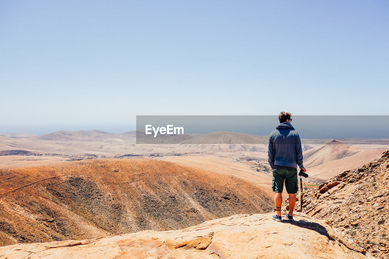 Rear view of man standing on rock looking at landscape against blue sky