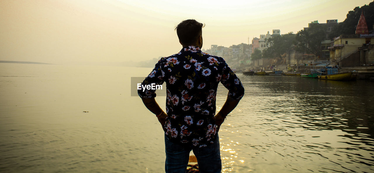 REAR VIEW OF MAN STANDING AT BEACH AGAINST SKY