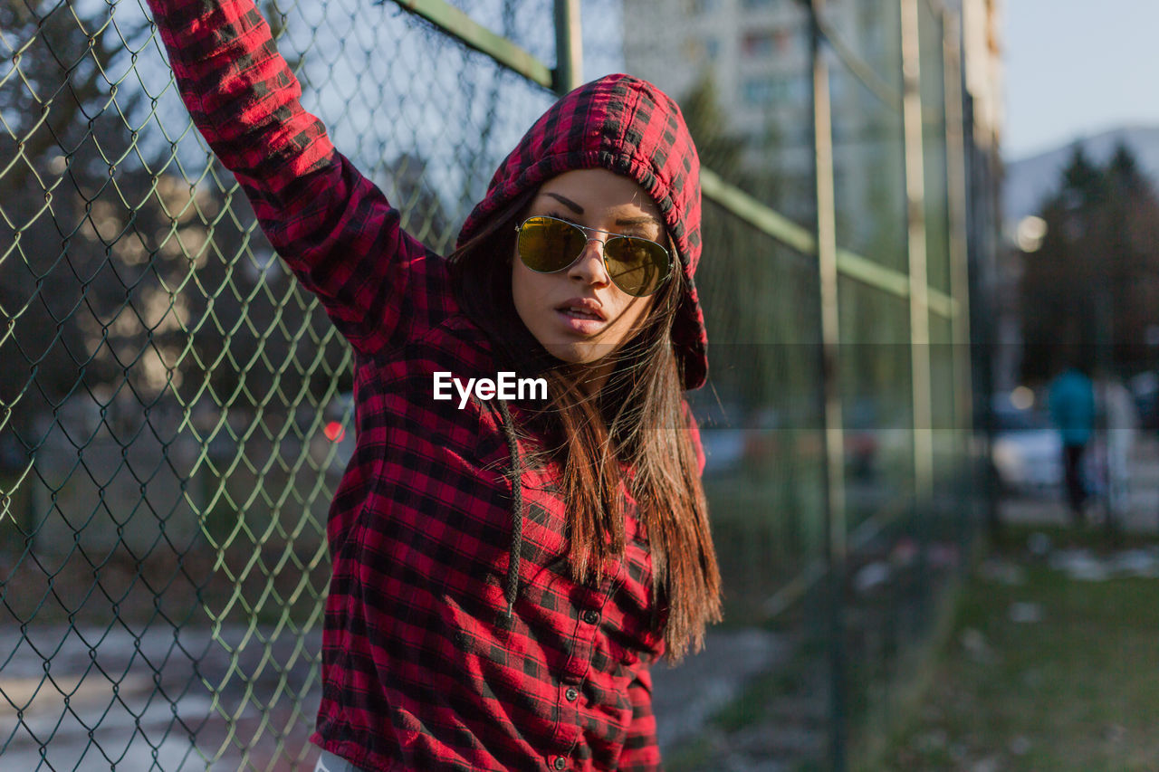 Close-up portrait of woman in sunglasses standing against fence