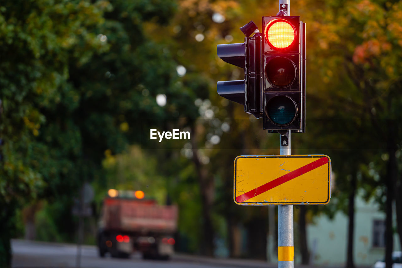 sign, road, road sign, transportation, stoplight, communication, guidance, traffic light, light, yellow, signaling device, lighting, city, street, road signal, red light, traffic, light fixture, traffic sign, mode of transportation, tree, no people, focus on foreground, warning sign, illuminated, red, nature, outdoors, symbol, vehicle, lighting equipment, day, plant, stop sign, city street, travel, signage, directional sign, car, motion, motor vehicle, city life