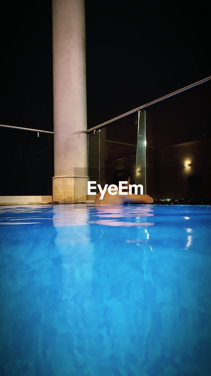 Shirtless man in blue swimming pool against clear sky at night