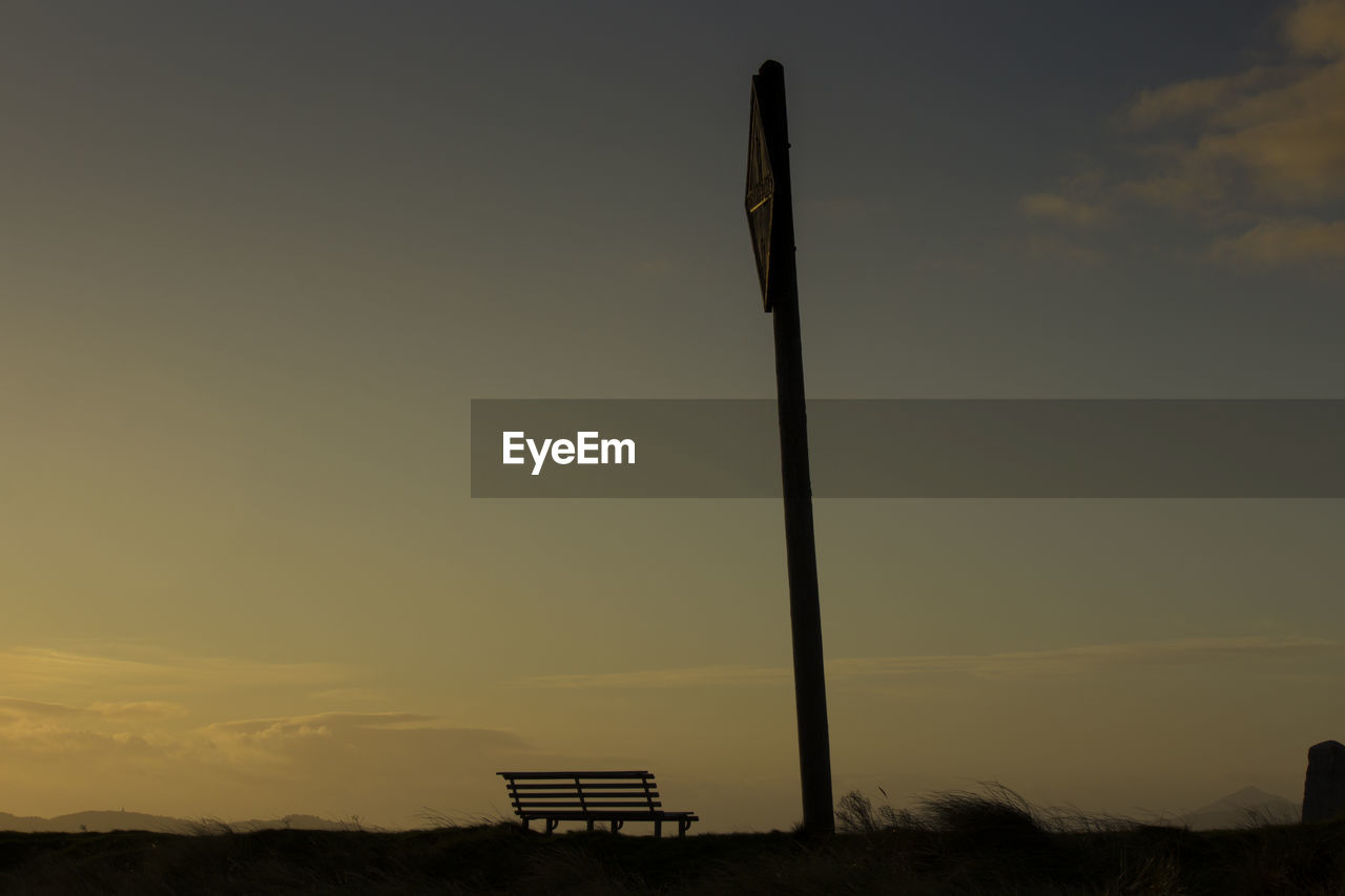 Silhouette pole and empty bench on field against sky at sunset