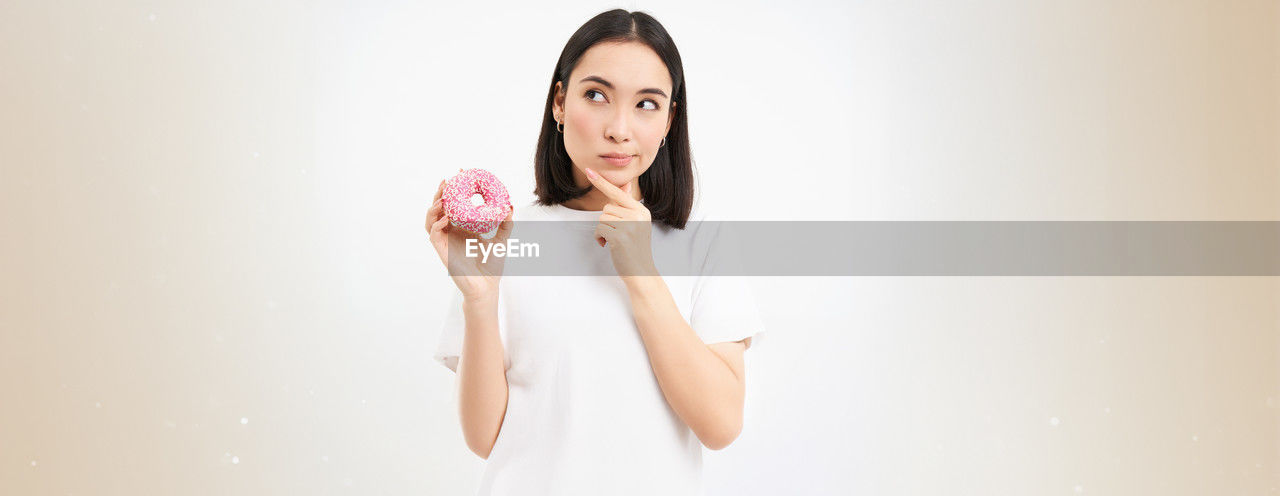 one person, women, adult, indoors, portrait, young adult, photo shoot, studio shot, hand, pink, clothing, looking at camera, long hair, waist up, copy space, finger, human face, hairstyle, person, emotion, skin, front view, lifestyles, holding, white background, food and drink, human leg, freshness, female, brown hair, smiling, happiness, fashion