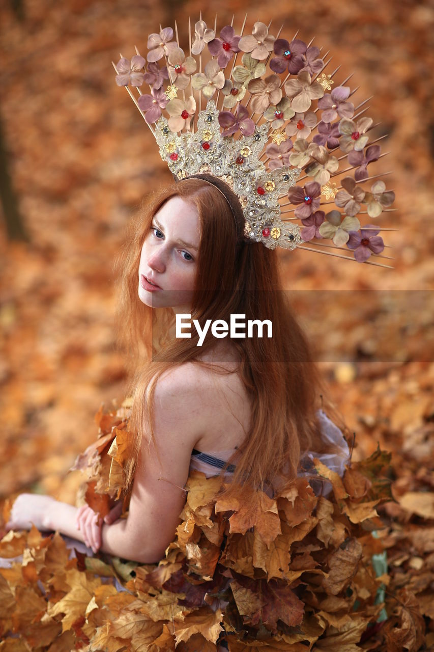 Thoughtful young woman covered with leaves sitting at forest during autumn