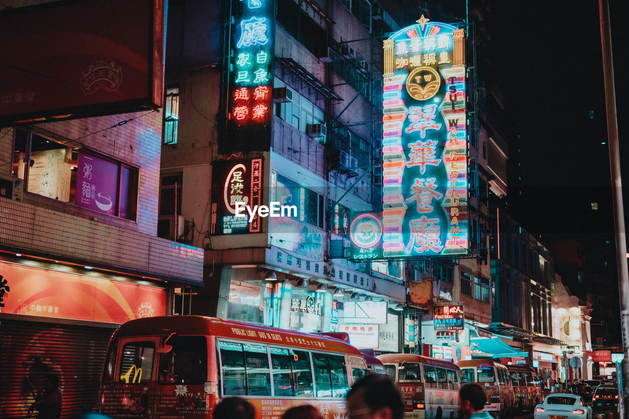 PEOPLE ON ILLUMINATED STREET AMIDST BUILDINGS IN CITY