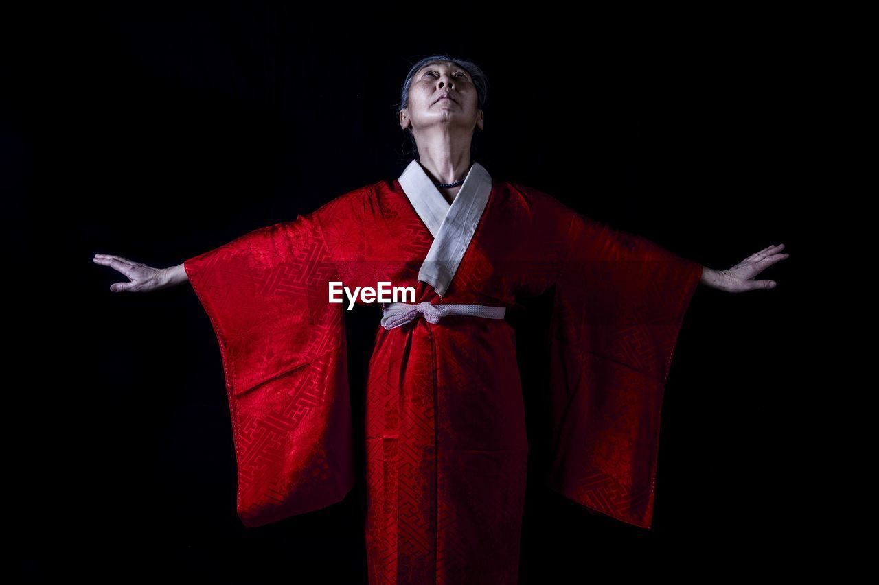 Thoughtful senior woman with arms outstretched standing in red kimono against black background