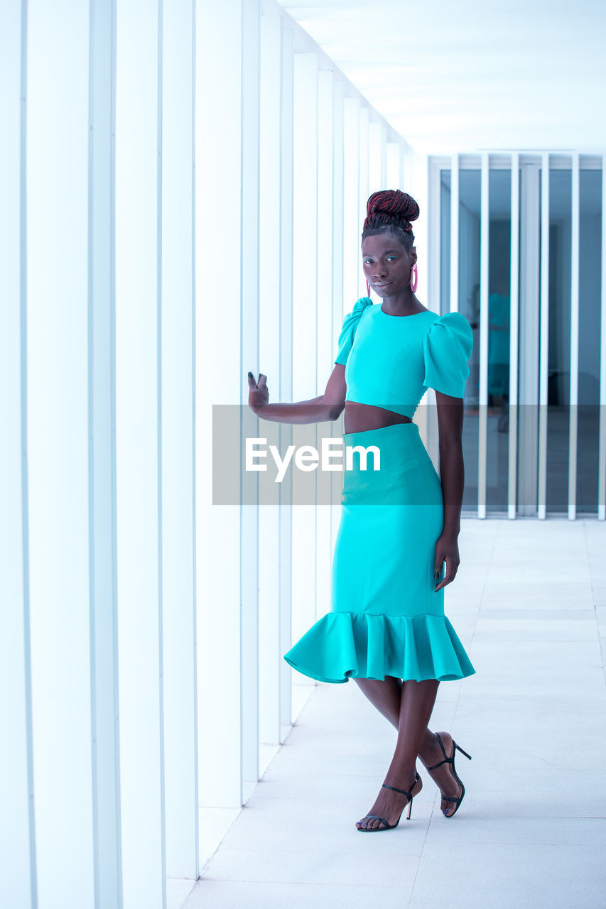 FULL LENGTH PORTRAIT OF SMILING WOMAN STANDING AGAINST CORRIDOR