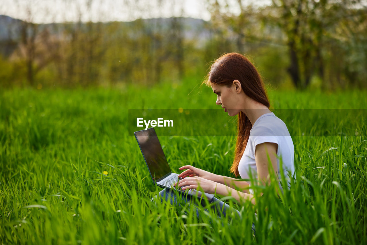 young woman using laptop while sitting on field