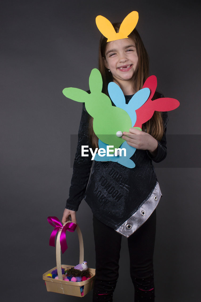 Portrait of smiling girl with eater bunny and pom pom in basket against gray background