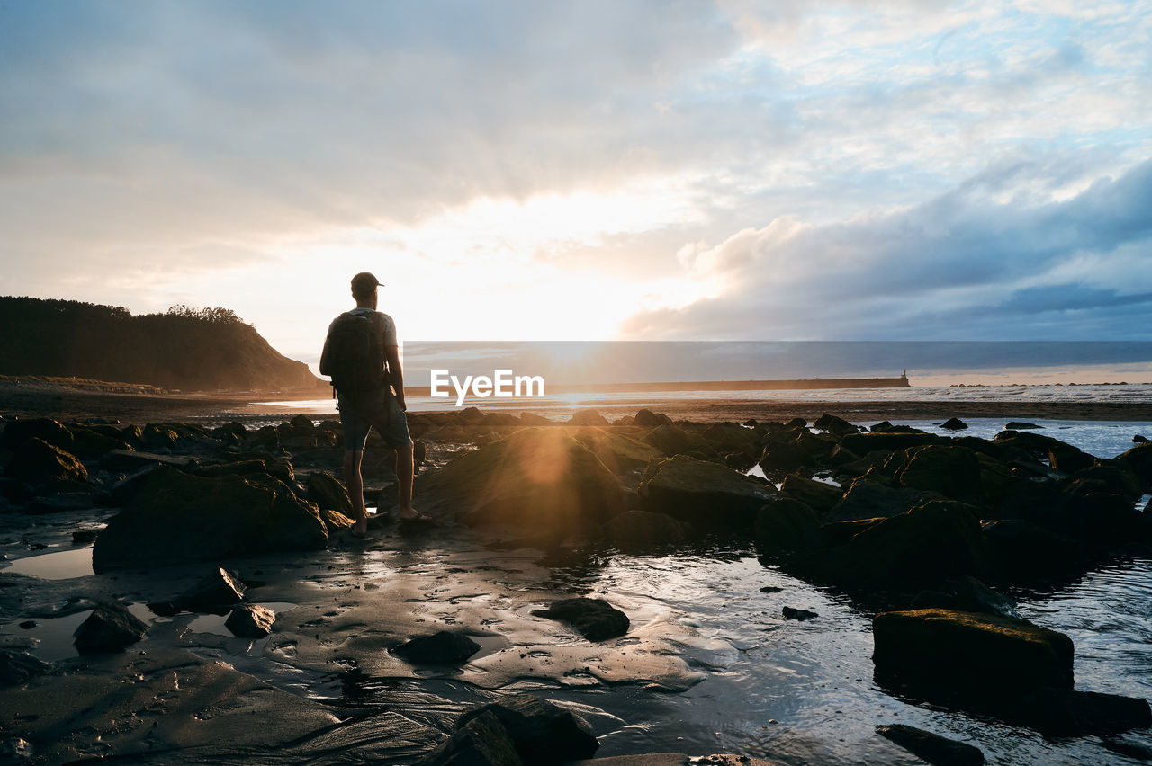 Back view full body of unrecognizable male tourist with backpack standing on boulders of wet coast and enjoying view of sea