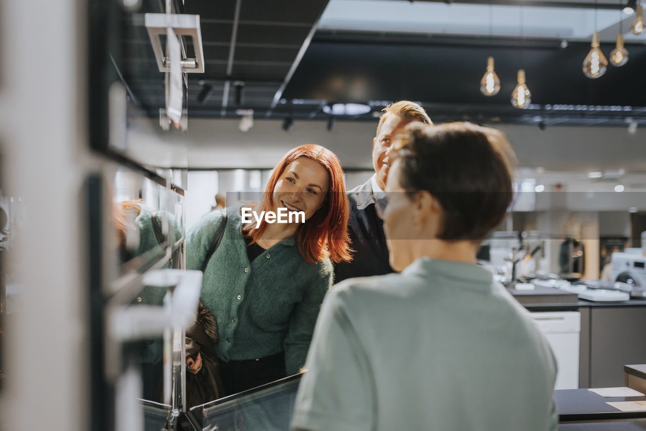 Happy couple looking at microwave oven while saleswoman assisting at electronics store