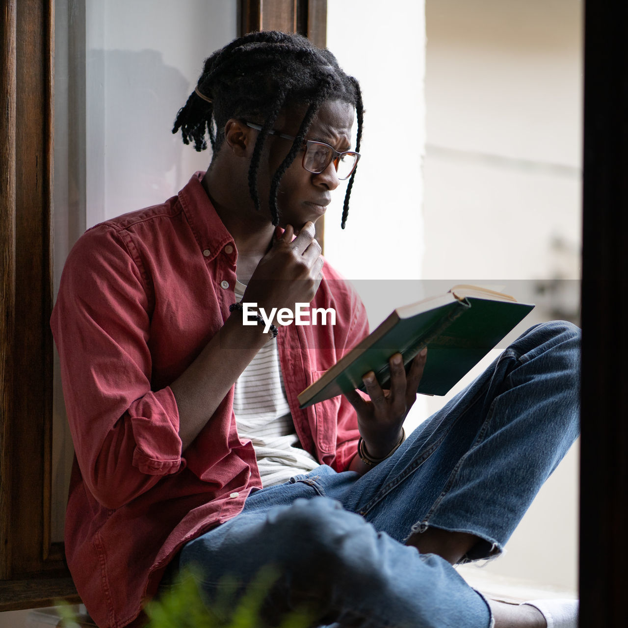 Concentrated inquisitive african american man student reading book sits on windowsill near window