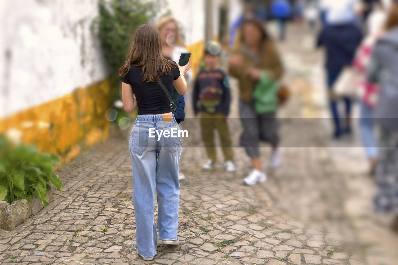 women, casual clothing, adult, city, architecture, footpath, full length, one person, jeans, street, young adult, female, lifestyles, walking, blond hair, day, spring, city life, hairstyle, rear view, child, outdoors, leisure activity, emotion, cobblestone, focus on foreground, sidewalk, motion, person, long hair, childhood, clothing, nature, standing, looking, selective focus